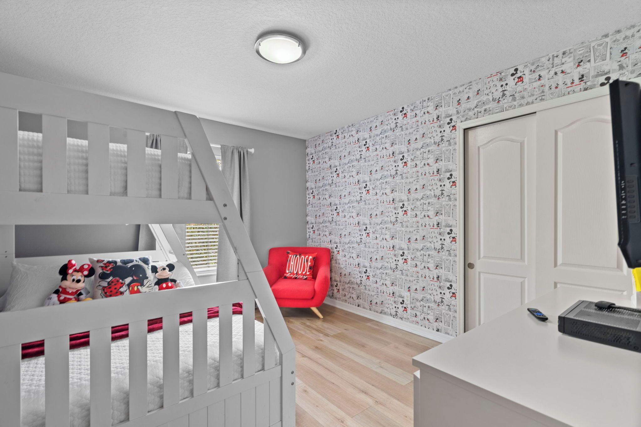 A child's bedroom with a bunk bed, a red chair, a white door, and a wall with cartoon character wallpaper.