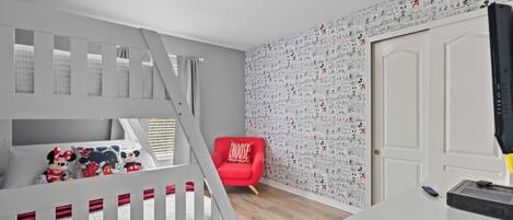 A child's bedroom with a bunk bed, a red chair, a white door, and a wall with cartoon character wallpaper.