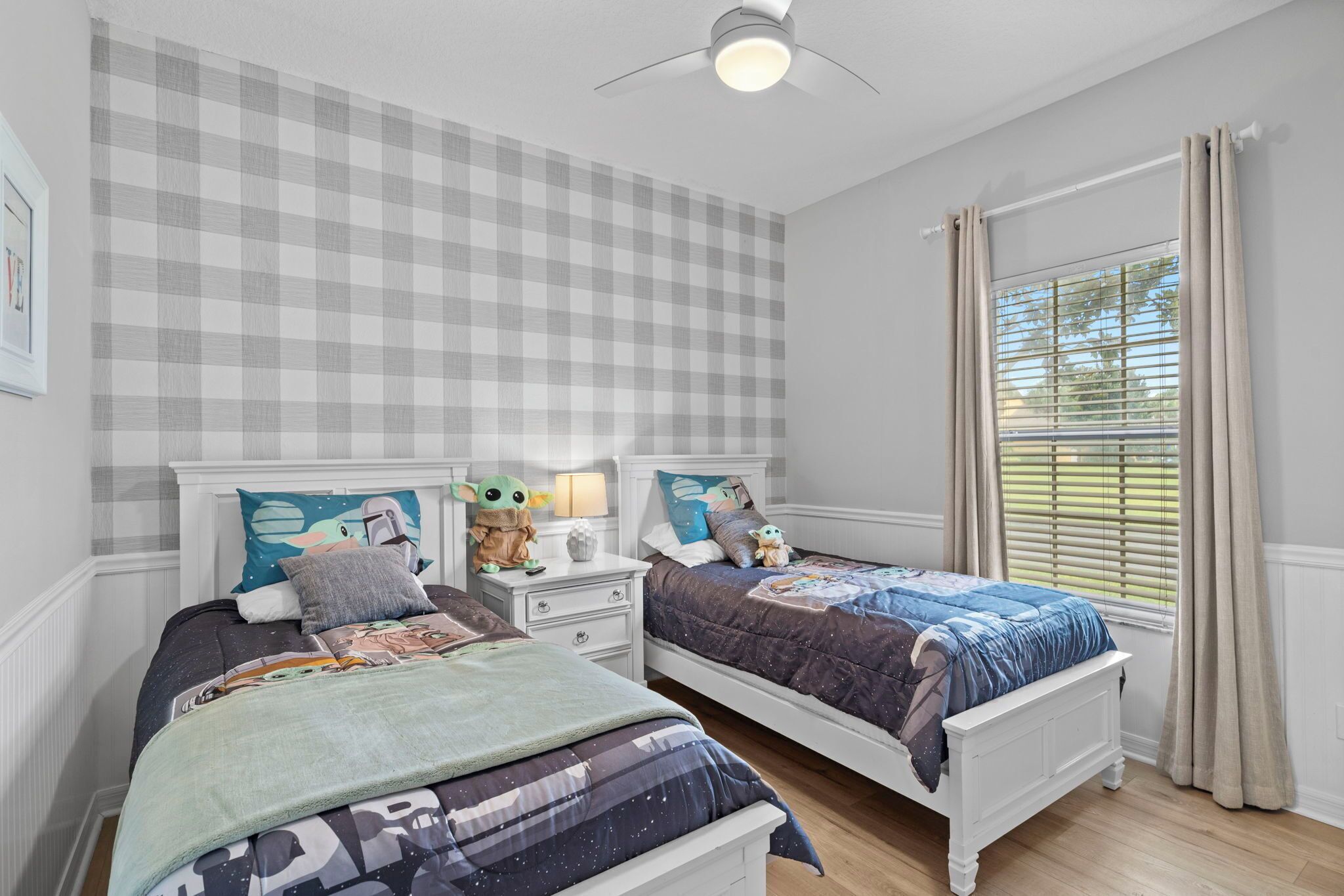 A bedroom with two beds, checkered wallpaper, a ceiling fan, and a window with curtains.