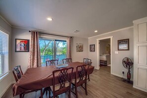 Uma sala de jantar com uma mesa, cadeiras, uma janela com cortinas e vista para o exterior arborizado.