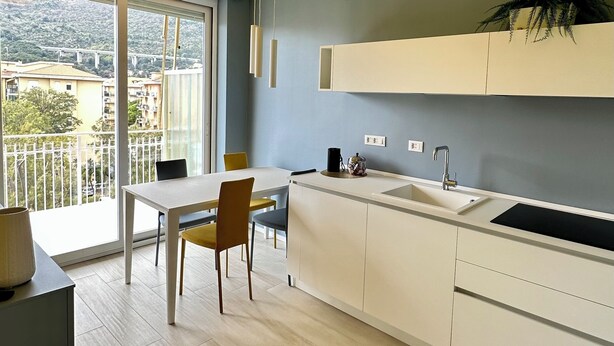 A modern kitchen with a large window, a dining area, and a balcony with a view of buildings and greenery. - Delightful apartment with modern and comfortable style near the beach (Marina di Andora)