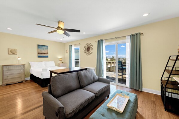 A living room with a gray sofa, a wooden coffee table, a ceiling fan, a bed with white bedding, and a view of the outdoors through large windows.