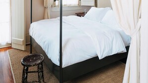A four-poster bed with white linens, a wooden stool, and a chandelier.