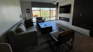 Ein modernes Wohnzimmer mit einer Couch, einem Couchtisch und Sesseln, einem Kamin und großen Fenstern mit Blick auf Gebäude und Bäume.