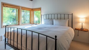 Une chambre à coucher avec un lit, deux fenêtres, une table de chevet avec une lampe et une étagère en bois.