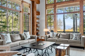 A spacious living room with two sofas, a coffee table, and a lamp. Large windows provide a view of trees and mountains.