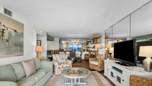 Un salón amplio con un sofá grande, una mesa de centro de vidrio y un televisor sobre un mueble blanco. Se aprecia un área de comedor al fondo con mobiliario de mimbre y vistas al exterior.