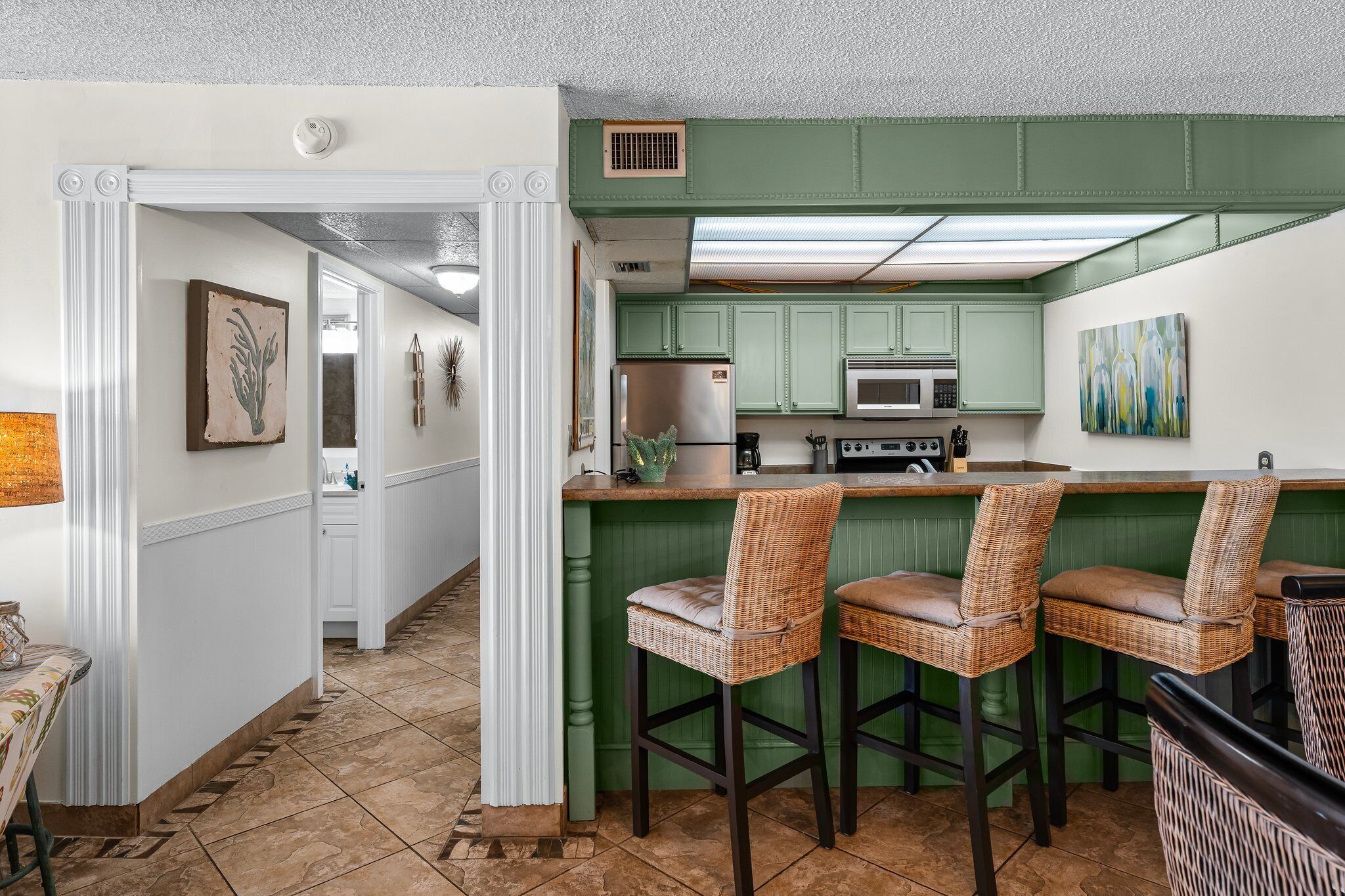 A kitchen with green cabinets, a stainless steel refrigerator, and a bar area with wicker chairs.