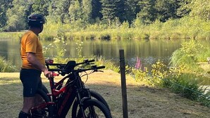 A person standing next to a mountain bike near a lake with a forest in the background.