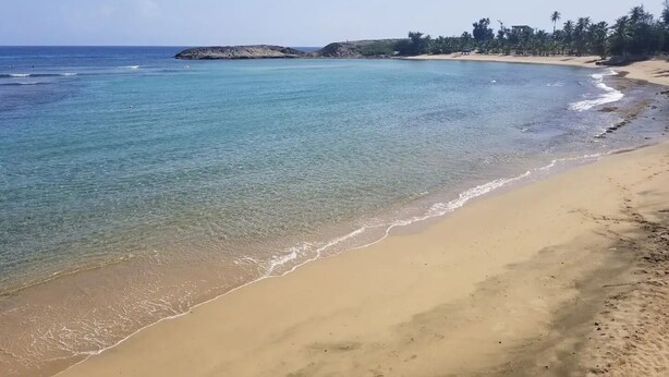 A beach with clear water, a sandy shore, and a rocky outcrop in the distance. - Waves & Chill 1 | Jobos Guest House (Isabela)