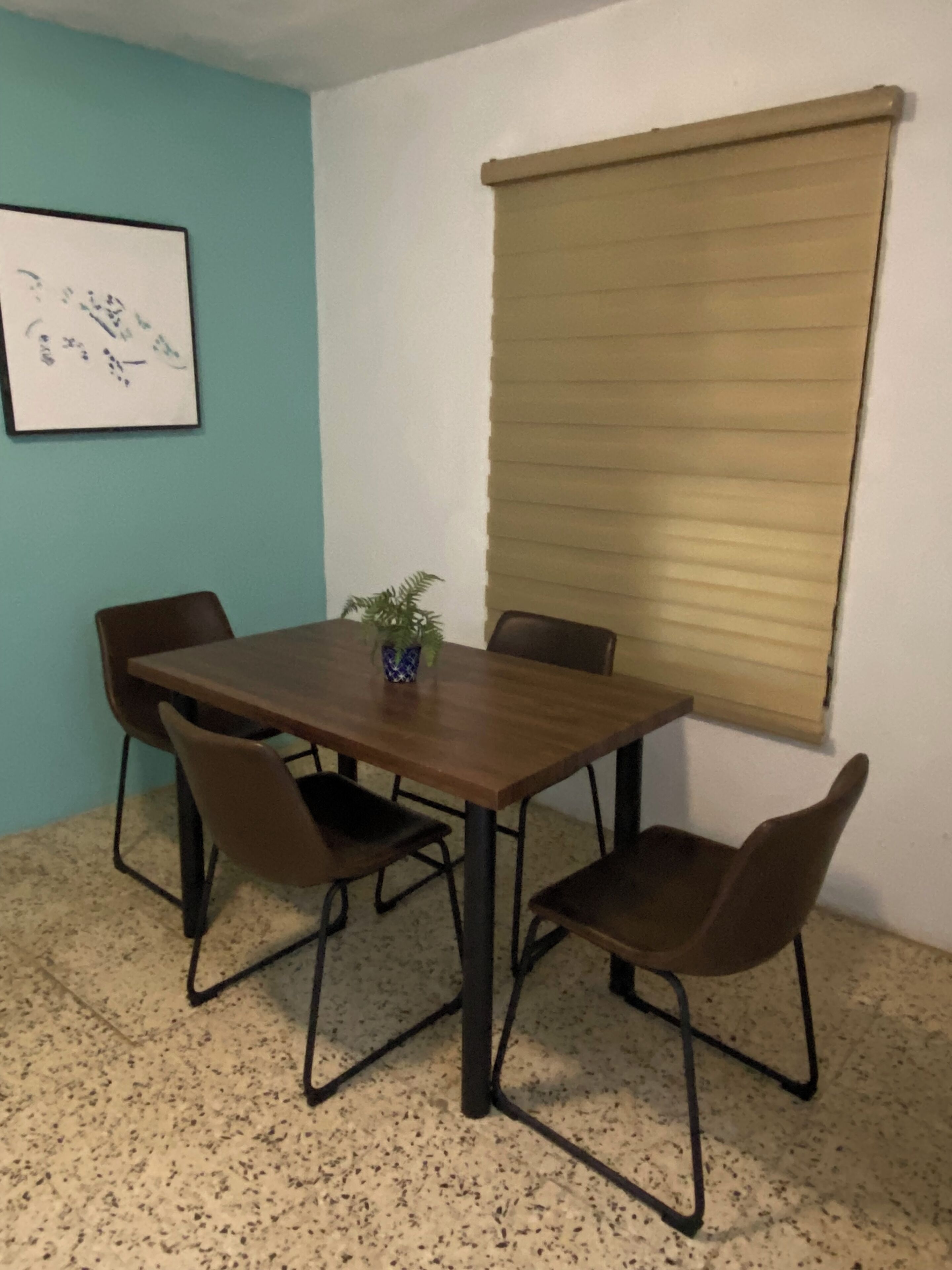 A room with a wooden table and four chairs, a potted plant, a framed picture on the wall, and a window with a beige blind.