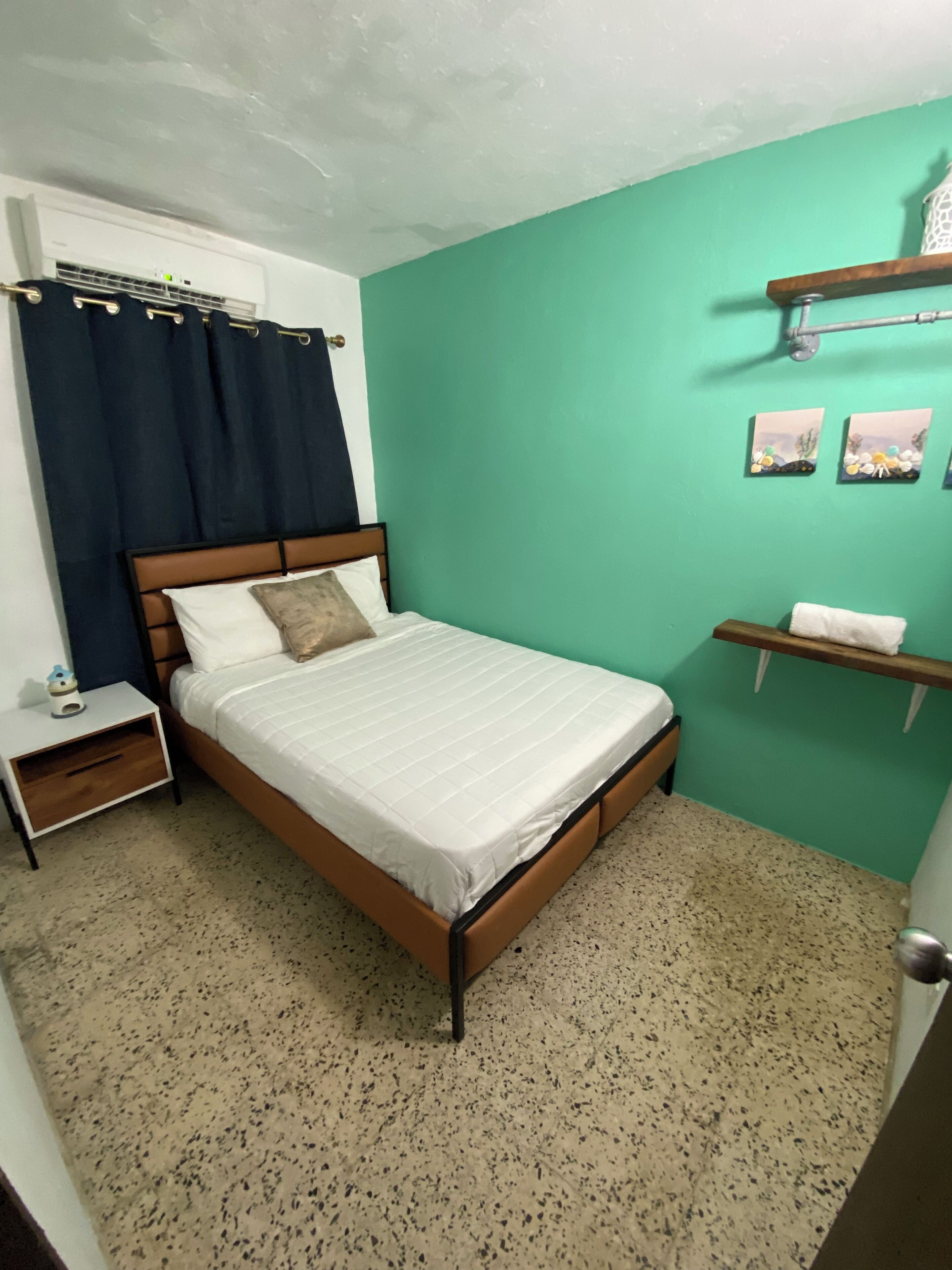 A bedroom with a bed, a nightstand, a wall-mounted shelf, and a window with curtains.