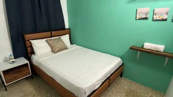 A bedroom with a bed, a nightstand, a wall-mounted shelf, and a window with curtains.