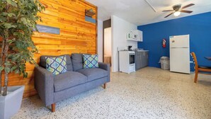 Un ambiente compatto con angolo cottura, un divano grigio con cuscini decorati, una parete a parete in legno e una ventola a soffitto.