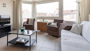 Un salon moderne doté d’un téléviseur à écran plat, d’une table basse en verre, de deux canapés, d’une commode en bois et d’une grande fenêtre offrant une vue sur la ville.