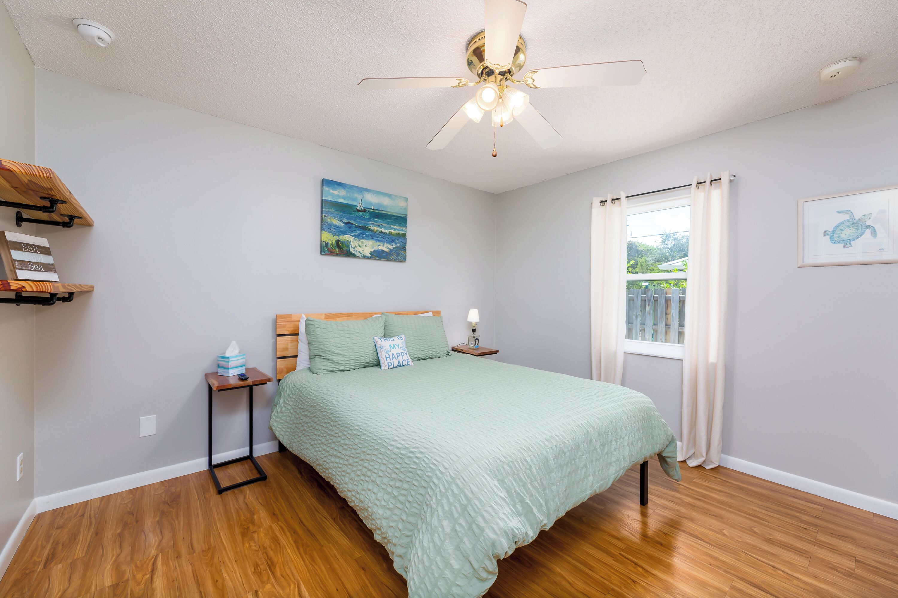 A bedroom with a bed, a ceiling fan, a nightstand, a window with curtains, and a wall with a painting.