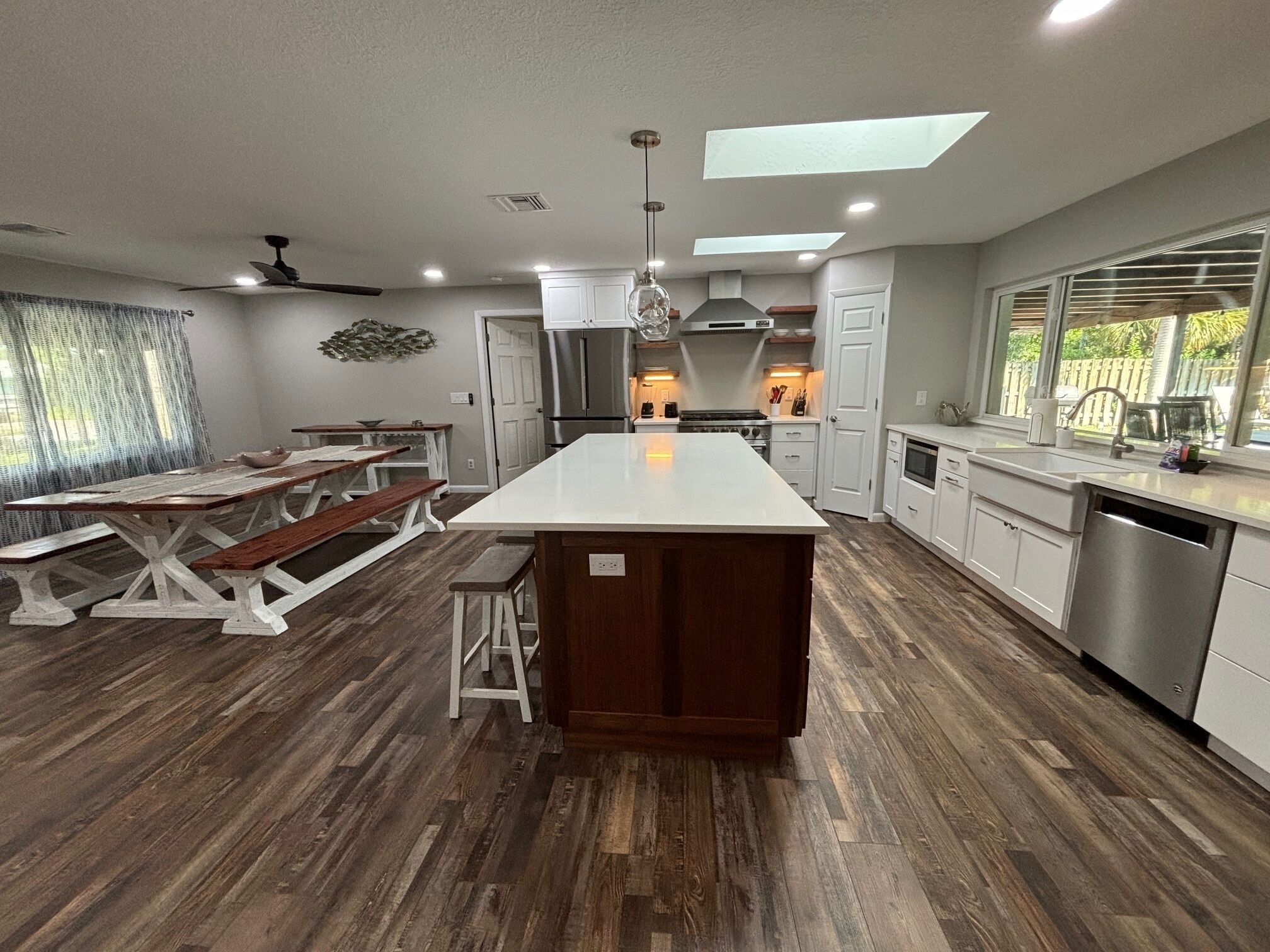 A modern kitchen with a central island, wooden flooring, white cabinetry, and a large window providing natural light.