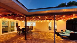Un patio al aire libre con piso de madera, zona de estar cubierta y luces colgantes.