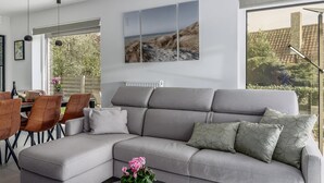 A modern living room with a grey sectional sofa, a dining area with wooden chairs, and a large window with a view of greenery and a building. - 2 bedroom accommodation in Oostduinkerke (Oostduinkerke)