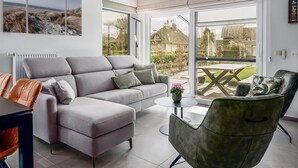 A modern living room with a grey sofa, a green armchair, and a small round table. There is a large window with a view of a garden and a house. - 2 bedroom accommodation in Oostduinkerke (Oostduinkerke)