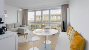 En moderne stue med sofa, spisebord med stole og et stort vindue der giver udsigt til bygninger.