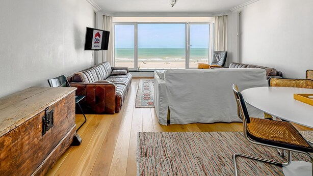 A modern hotel room with a large window offering a beach view, a sofa, a dining table, and a wooden chest of drawers. - 2 bedroom accommodation in Sint Idesbald (Sint Idesbald)