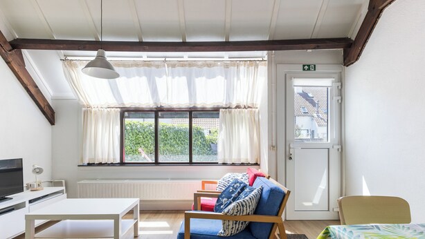 A room with a wooden-framed ceiling, a white table, a blue sofa with patterned cushions, a television, and a window with white curtains. - 2 bedroom accommodation in Nieuwpoort (Nieuwpoort)