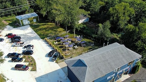 Vista aérea de un edificio con techo azul y la inscripción 'BEHR BREWING' en el lateral. Se observa un estacionamiento con varios autos y un área verde con mesas y sombrillas.