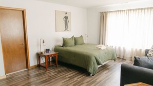 Habitación de hotel con suelo de madera, cama con ropa de cama verde, mesita de noche con lámpara, una silla y un cuadro en la pared.