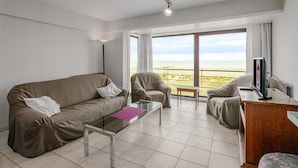 A modern hotel room with a large balcony, a sofa, a chair, a small table, and a TV.