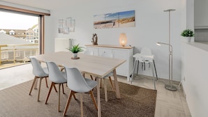 A modern kitchen with a dining area, featuring a large table, chairs, a plant, a lamp, and a view of buildings outside.