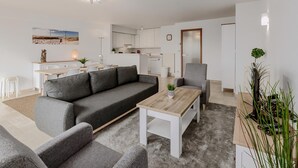 A modern living room with a grey sofa, a wooden coffee table, and a grey armchair. There is a kitchen in the background with white cabinets and a dining area with a table and chairs.