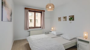 A bedroom with two beds, a window with curtains, a bedside table with a lamp, and wall-mounted artwork.