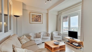 A modern living room with a beige sofa, two white armchairs, a wooden coffee table, a flat-screen TV, and a large window with curtains. - 2 bedroom accommodation in Koksijde (Koksijde)