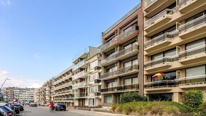 A multi-story residential building with balconies, a parking area, and a clear sky.