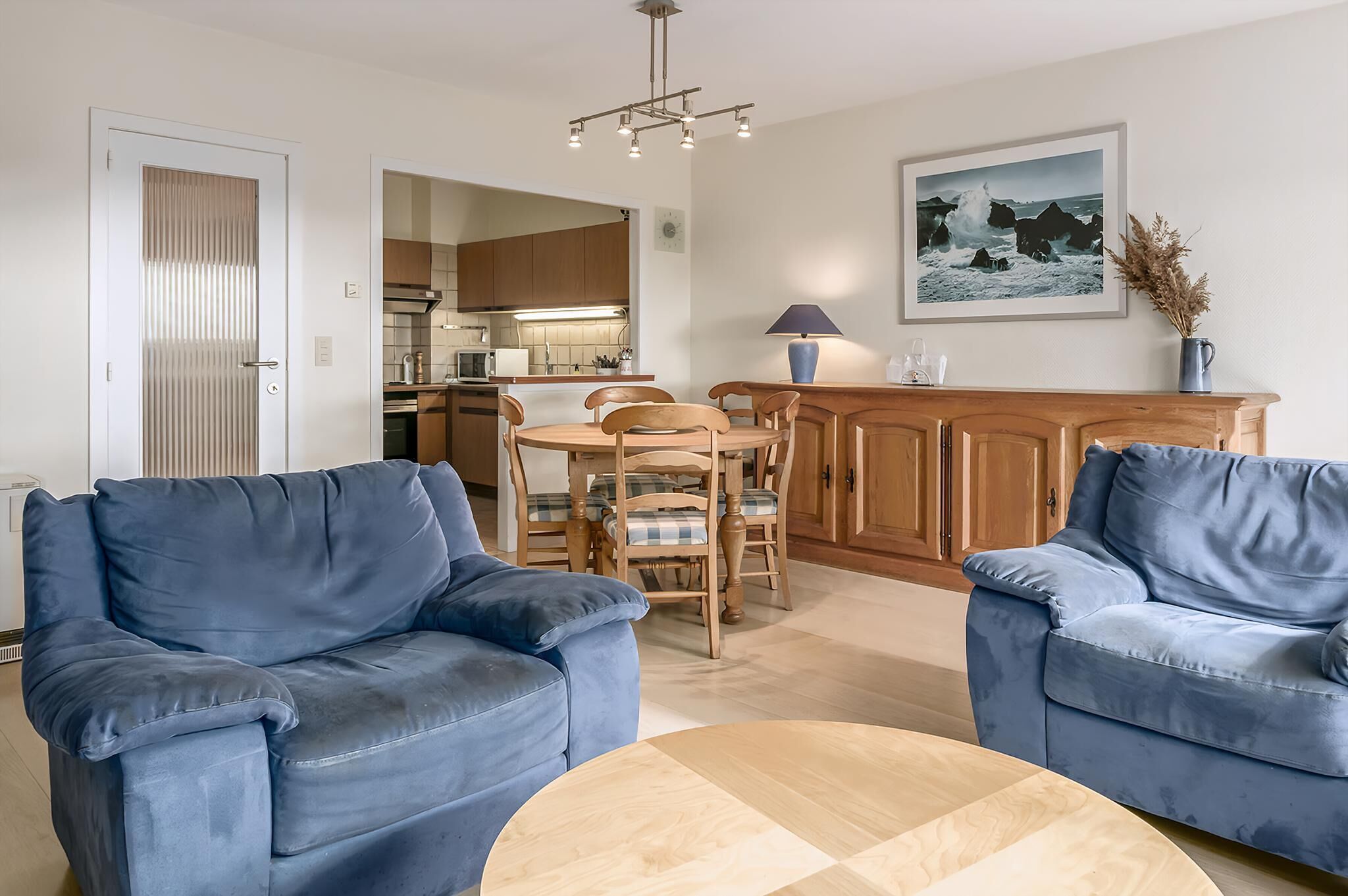 A living room with two blue sofas, a wooden dining table with chairs, a kitchen in the background, and a framed picture on the wall.