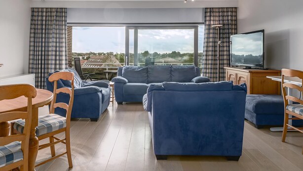 A modern living room with a blue sofa set, a wooden dining table with chairs, and a television on a wooden cabinet.