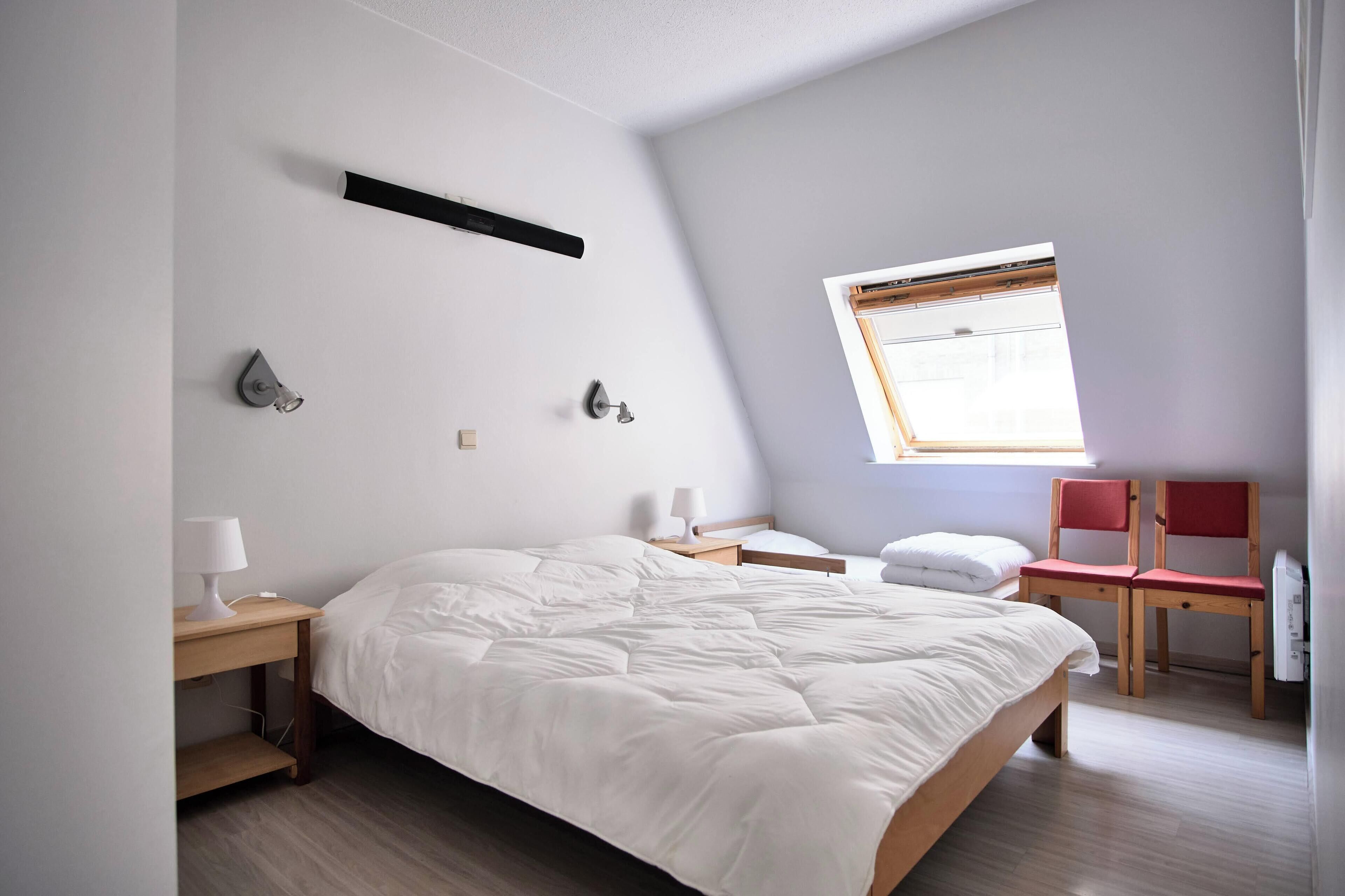A bedroom with a large bed, bedside tables with lamps, a window with blinds, and a red chair.