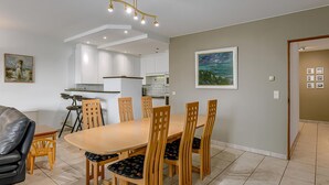 A modern dining area with a wooden table and chairs, a small kitchen in the background, and artwork on the walls.