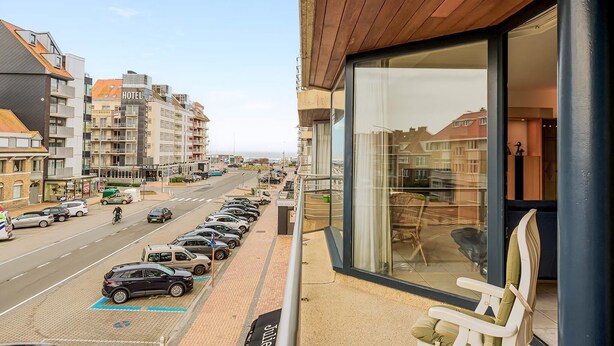 A hotel with a balcony overlooking a street with parked cars and a view of the sea.