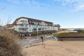 Um edifício à beira-mar com várias varandas, uma praia arenosa em primeiro plano e um céu limpo.