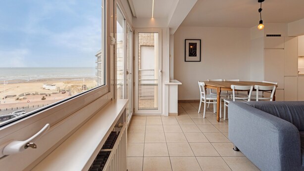 A modern living room with a large window overlooking a beach, a dining area with a table and chairs, and a gray sofa. - 2 bedroom accommodation in Koksijde (Koksijde)