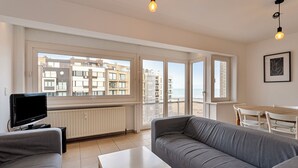 A modern living room with a grey sofa, a white coffee table, and a TV. Large windows offer a view of the cityscape. - 2 bedroom accommodation in Koksijde (Koksijde)