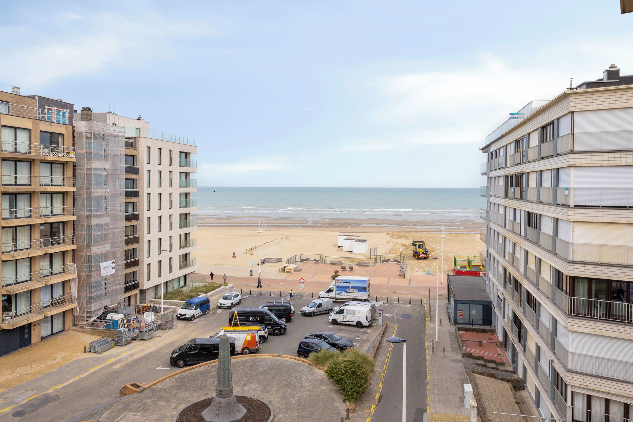 A coastal view with buildings, a beach, and parked vehicles.