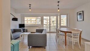 A modern living room with a sofa, a coffee table, a TV, and a dining table with chairs. - 2 bedroom accommodation in Koksijde (Koksijde)