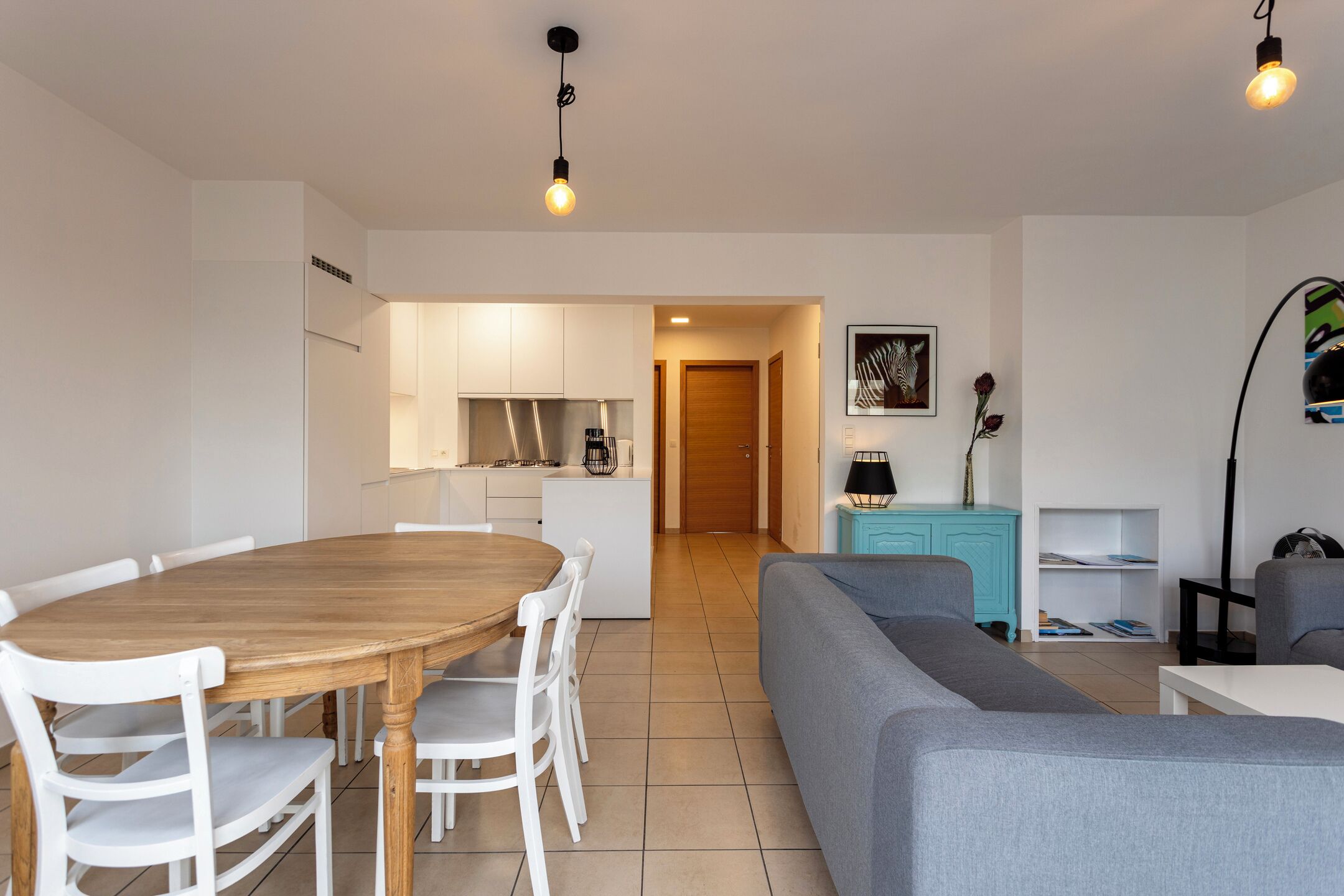 A modern living room with a grey sofa, a wooden dining table with white chairs, a kitchen area with white cabinets, and a teal side table.