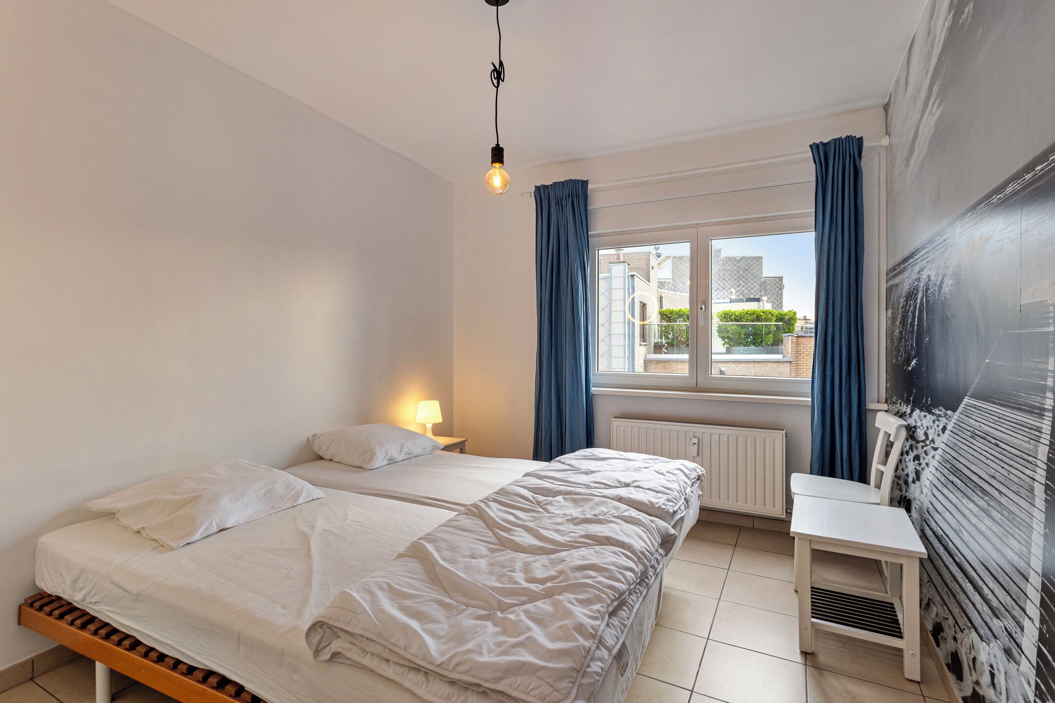 Quarto com duas camas, uma escrivaninha, uma janela com cortinas e um radiador.