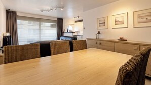 Un espace repas moderne avec une table en bois et des chaises en osier, une cuisine en arrière-plan et des tableaux encadrés au mur.