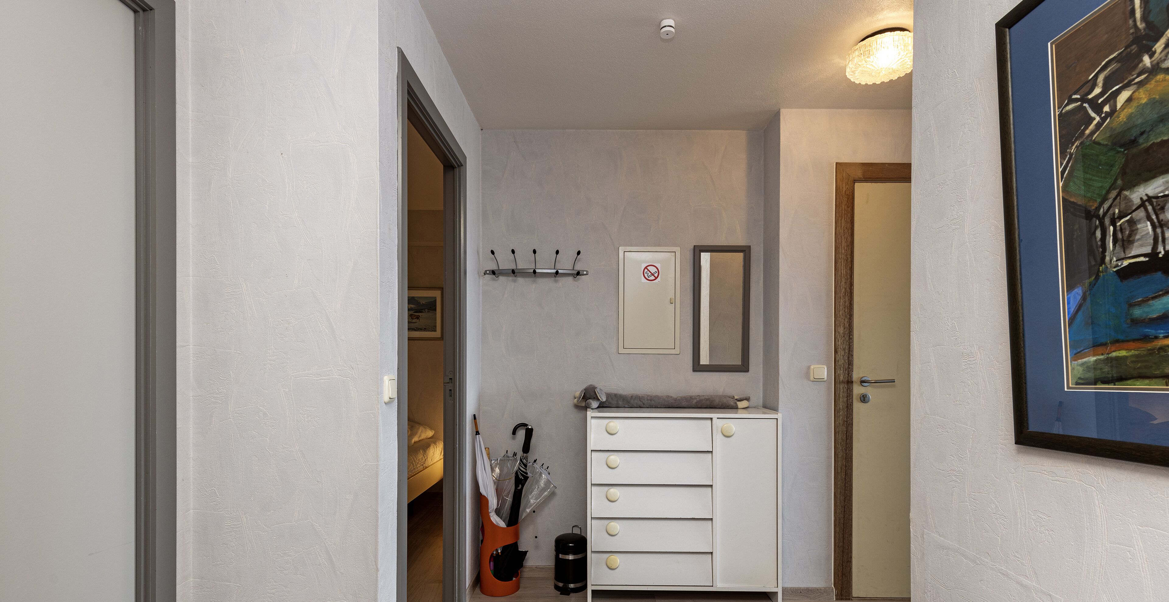 Un couloir avec un porte-manteau, une porte sur laquelle est apposé un panneau « interdiction de fumer », une petite armoire et un tableau encadré accroché au mur.
