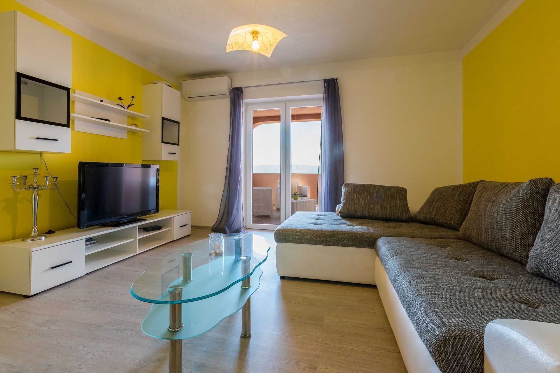 A modern living room with a glass coffee table, a sofa, a flat-screen TV, and a balcony with a view.